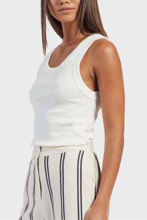 A woman is wearing the Academy Women Essential Rib Tank, matched perfectly with high-waisted, light-colored striped pants. Her long, straight hair cascades down as she poses slightly turned to the side, highlighting her casual yet stylish summer ensemble.