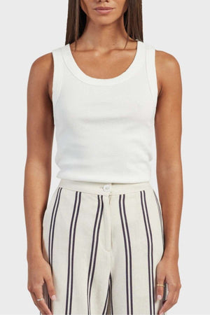 A person wearing an Academy Women Essential Rib Tank, which is a white sleeveless premium garment washed rib top, and high-waisted cream-colored pants featuring vertical navy and white stripes. They have long straight hair with their hands relaxed at their sides, embodying the ideal summer outfit. The background is a plain light gray.
