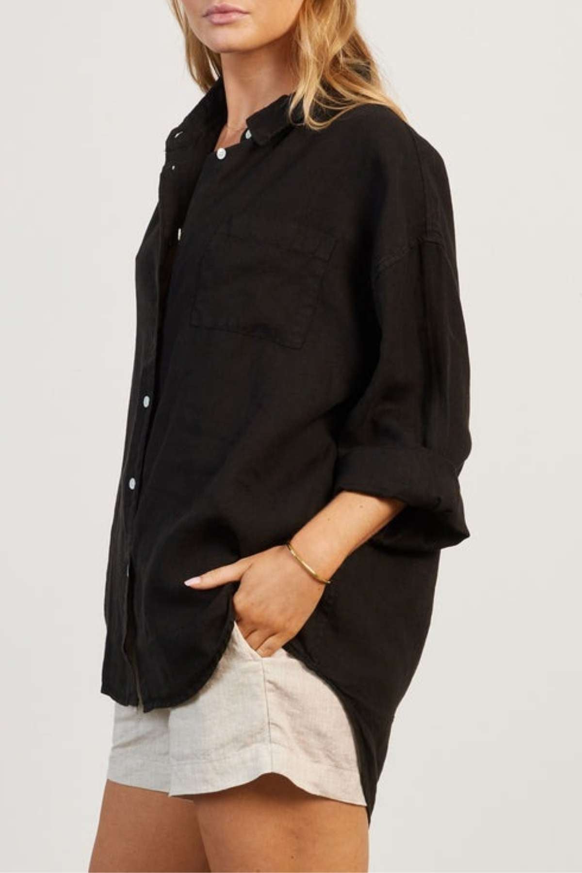 A person with long, blonde hair is wearing the Academy Women Hampton Linen Shirt— a loose, garment-dyed black button-up shirt featuring mother-of-pearl buttons— with rolled-up sleeves and beige shorts. With one hand in their pocket, they stand against a plain background, their face partially visible and turned slightly to the side.