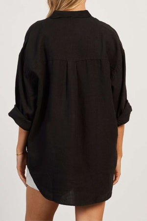 A person with long hair is shown from the back, wearing a black, loose-fitting, long-sleeve Academy Women Hampton Linen Shirt with the sleeves rolled up. They are also wearing light-colored shorts. The background is neutral.