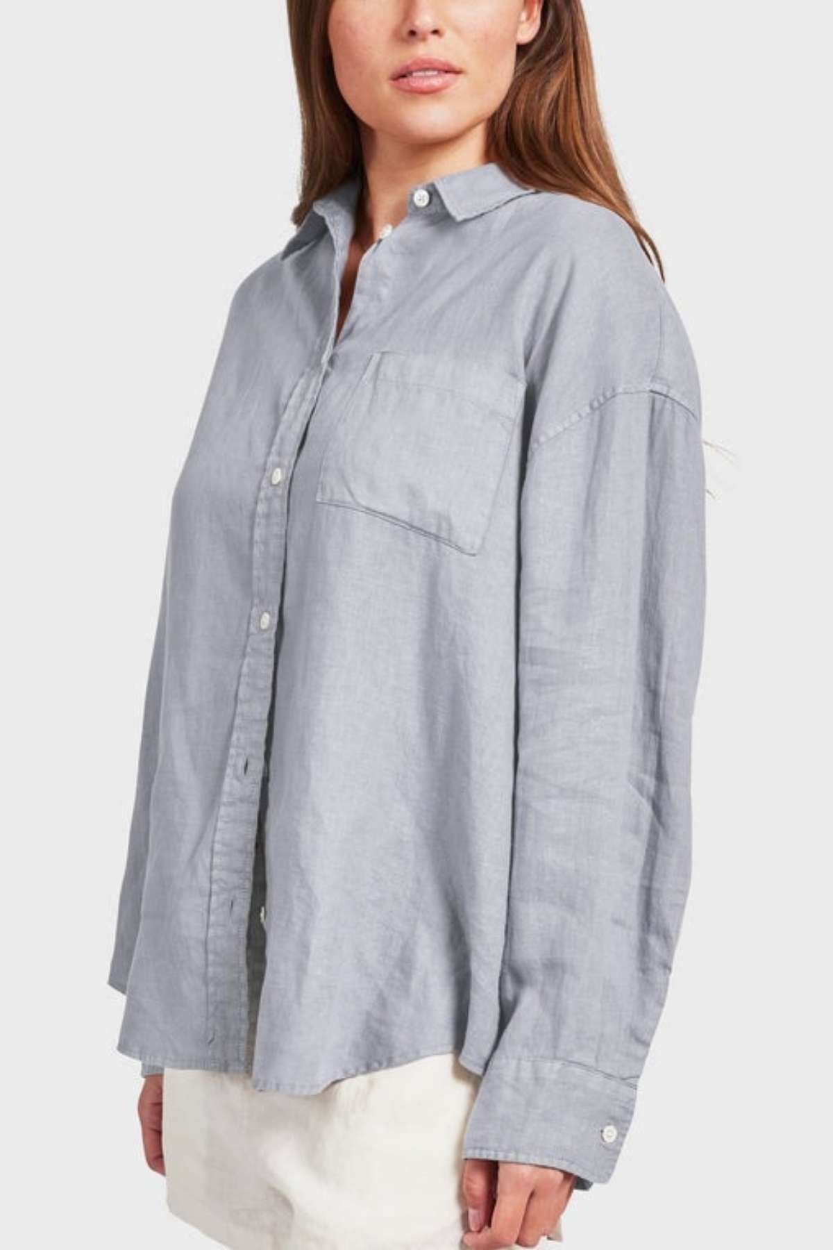 A person wearing an Academy Women Hampton Linen Shirt in light gray with a front pocket stands against a plain, light gray background. The garment-dyed shirt, celebrated for its extra softness, looks relaxed and comfortable even though the person's hands are not visible.