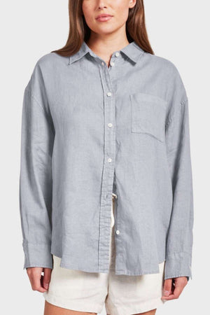 A person with long hair is wearing a light gray Academy Women Hampton Linen Shirt, celebrated for its exceptional softness. This garment-dyed shirt boasts long sleeves and a front pocket. Complementing their outfit are light-colored shorts, set against a plain, neutral background.