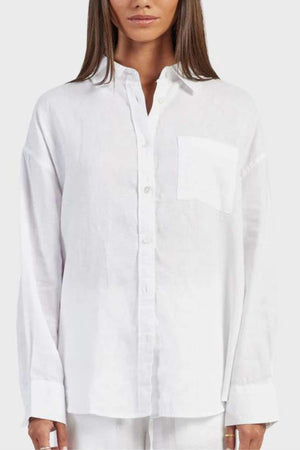 A person with long brown hair is wearing an Academy Women Hampton Linen Shirt in white, showcasing its relaxed fit and front pocket. The garment-dyed shirt is adorned with natural mother-of-pearl buttons and captured from mid-torso up against a neutral background.