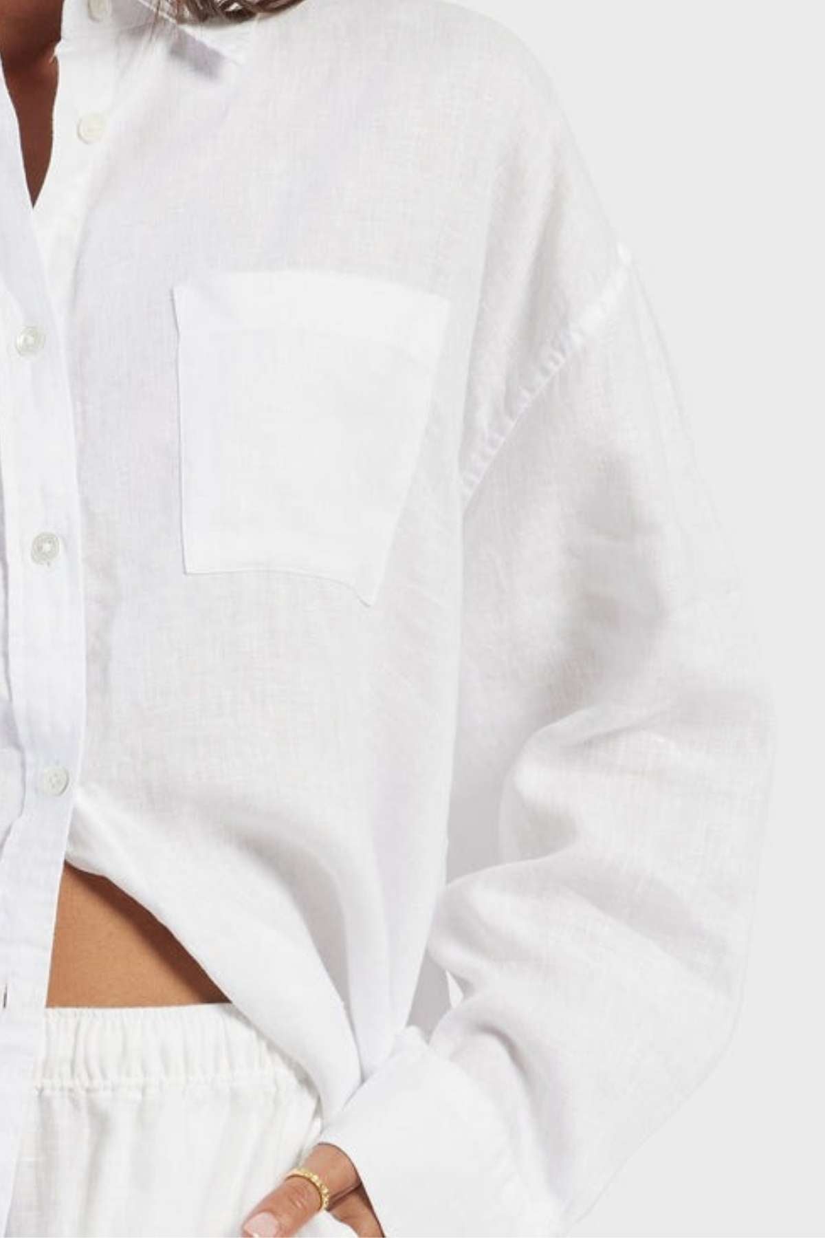 A person wearing a loose-fitting, white Academy Women Hampton Linen Shirt with natural mother-of-pearl buttons and cuffed long sleeves. The garment dyed shirt is slightly unbuttoned at the bottom, revealing matching white pants with an elastic waistband. The individual's hand rests at their hip, adorned with a gold ring.