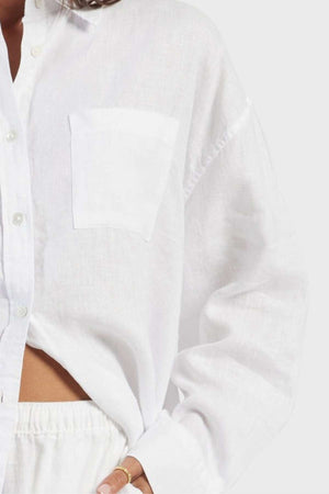 A person wearing a loose-fitting, white Academy Women Hampton Linen Shirt with natural mother-of-pearl buttons and cuffed long sleeves. The garment dyed shirt is slightly unbuttoned at the bottom, revealing matching white pants with an elastic waistband. The individual's hand rests at their hip, adorned with a gold ring.