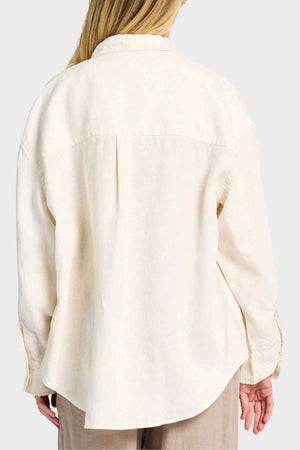 A person with long hair is seen from behind wearing a cream-colored Academy Women Hampton Linen Shirt, which features mother-of-pearl buttons. The shirt offers a loose fit and pairs perfectly with beige pants against a plain white background.