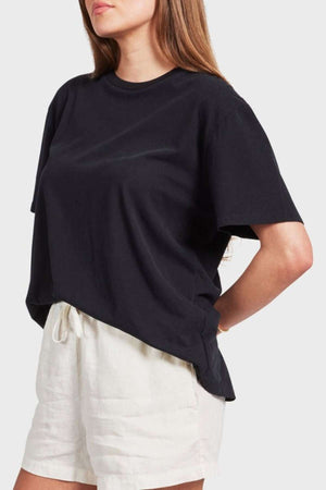 A person with long hair is wearing a loose-fitting, oversized *Academy Women Jimmy Boyfriend Tee* in black and light-colored drawstring shorts. The person is standing with a slight side profile and has their hands resting on their hips. The background is plain and light gray.