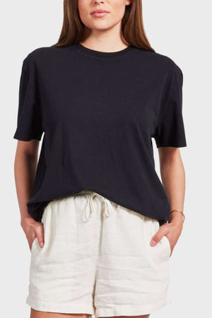 A person with long hair is wearing an oversized Academy Women Jimmy Boyfriend Tee in plain black and light-colored drawstring shorts. They have their hands tucked into the shorts' pockets and a gold bracelet on their left wrist. The background is plain and gray.