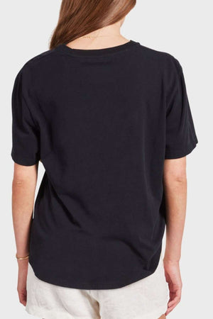 A person with long hair is shown from the back, wearing a black Academy Women Jimmy Boyfriend Tee and light-colored shorts. They have a gold bracelet on their left wrist. The background is plain and light-colored.