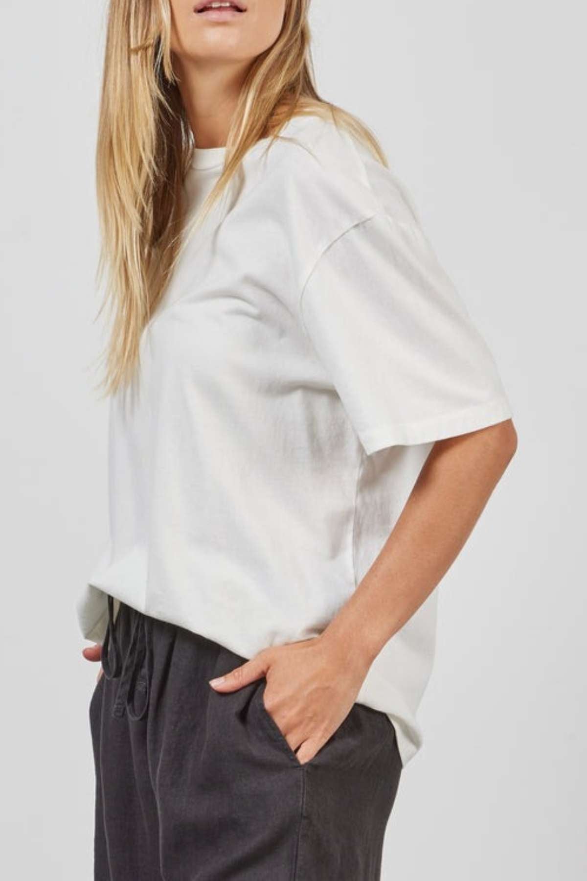 A person with long blonde hair is wearing an oversized Academy Women Jimmy Boyfriend Tee and dark pants. They have their right hand in their pocket and are standing in front of a plain, light-colored background. Their face is partially out of the frame.