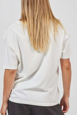 Rear view of an individual with long blonde hair, dressed in a loose-fitting Academy Women Jimmy Boyfriend Tee and dark pants. The person's right hand is partially visible, with fingers slightly bent, while they are turned away from the camera against a plain light background.