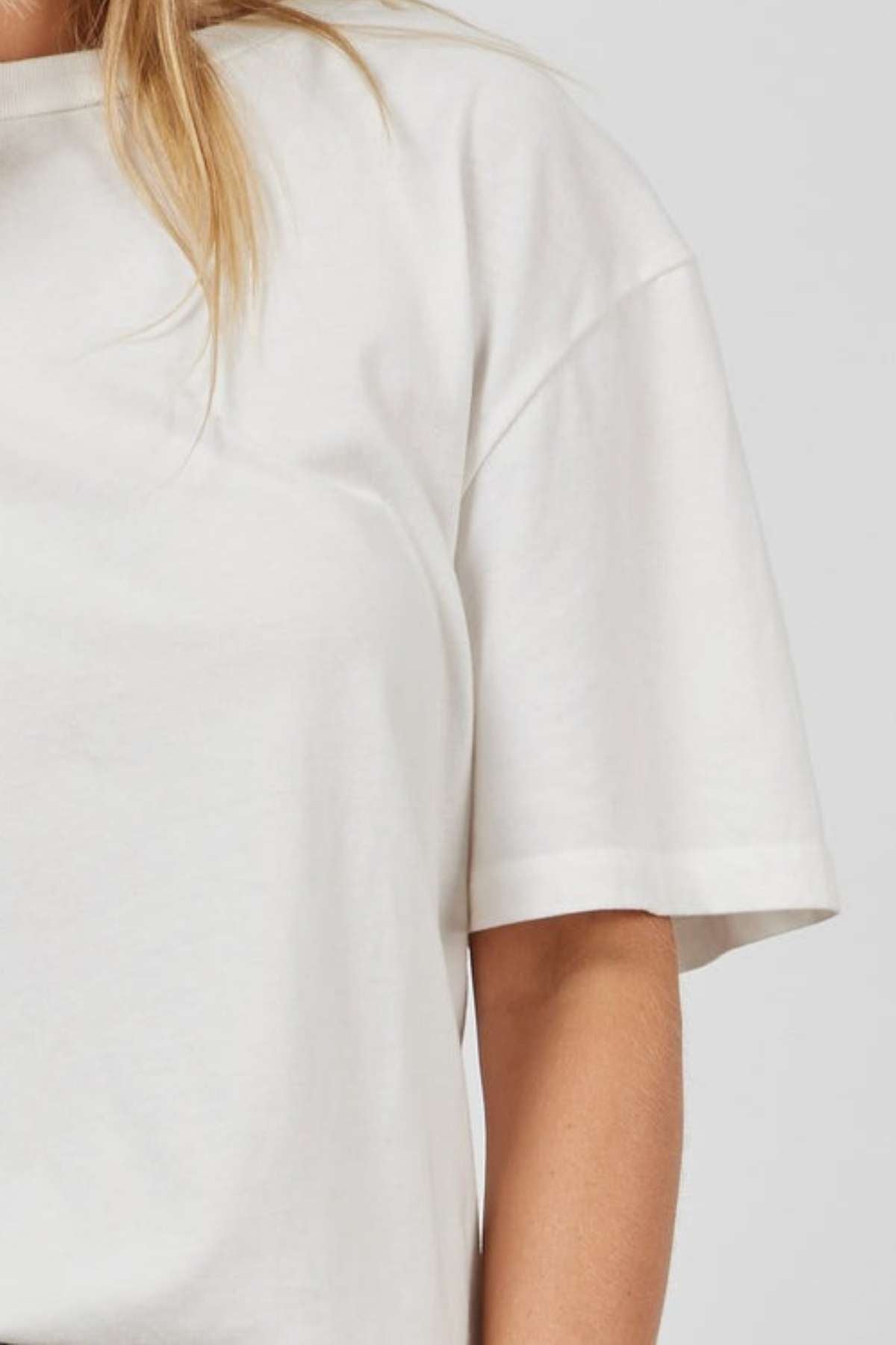 A close-up of a person wearing the Academy Women Jimmy Boyfriend Tee, an oversized plain white short-sleeve shirt. The image focuses on the shoulder, arm, and upper torso with blonde hair cascading over the left shoulder against a light gray background.