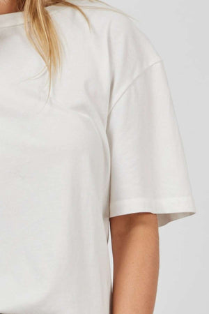 A close-up of a person wearing the Academy Women Jimmy Boyfriend Tee, an oversized plain white short-sleeve shirt. The image focuses on the shoulder, arm, and upper torso with blonde hair cascading over the left shoulder against a light gray background.