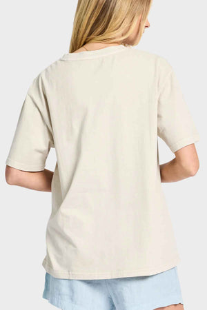 A person with long blonde hair stands with their back to the camera, wearing an oversized tee from the Academy Women Jimmy Boyfriend Tee line. The plain beige premium sanded jersey pairs effortlessly with light blue shorts against a simple, light gray background.