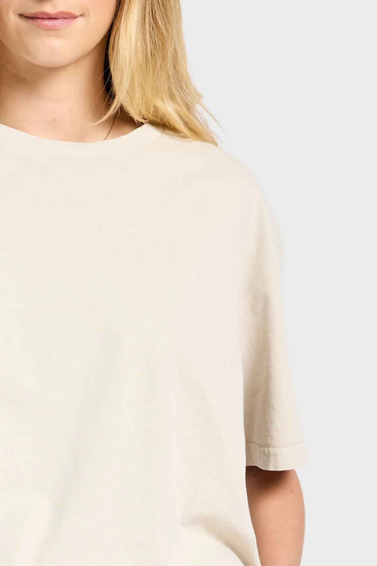 A person with long blonde hair is wearing an Academy Women Jimmy Boyfriend Tee in beige, crafted from premium sanded jersey. The image shows their upper torso and part of their face against a light gray background.