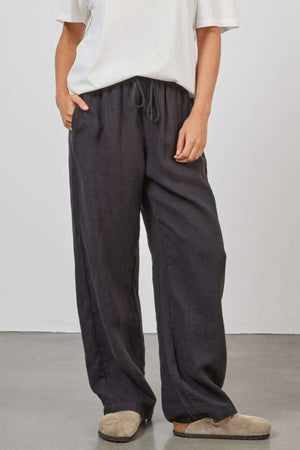 A person is wearing a white short-sleeve shirt and a pair of Academy Women Riviera Linen Pants, which are relaxed wide-leg black pants with a drawstring waist. They have one hand in their pocket and are standing on a plain, smooth surface. The person is also wearing light brown slippers.