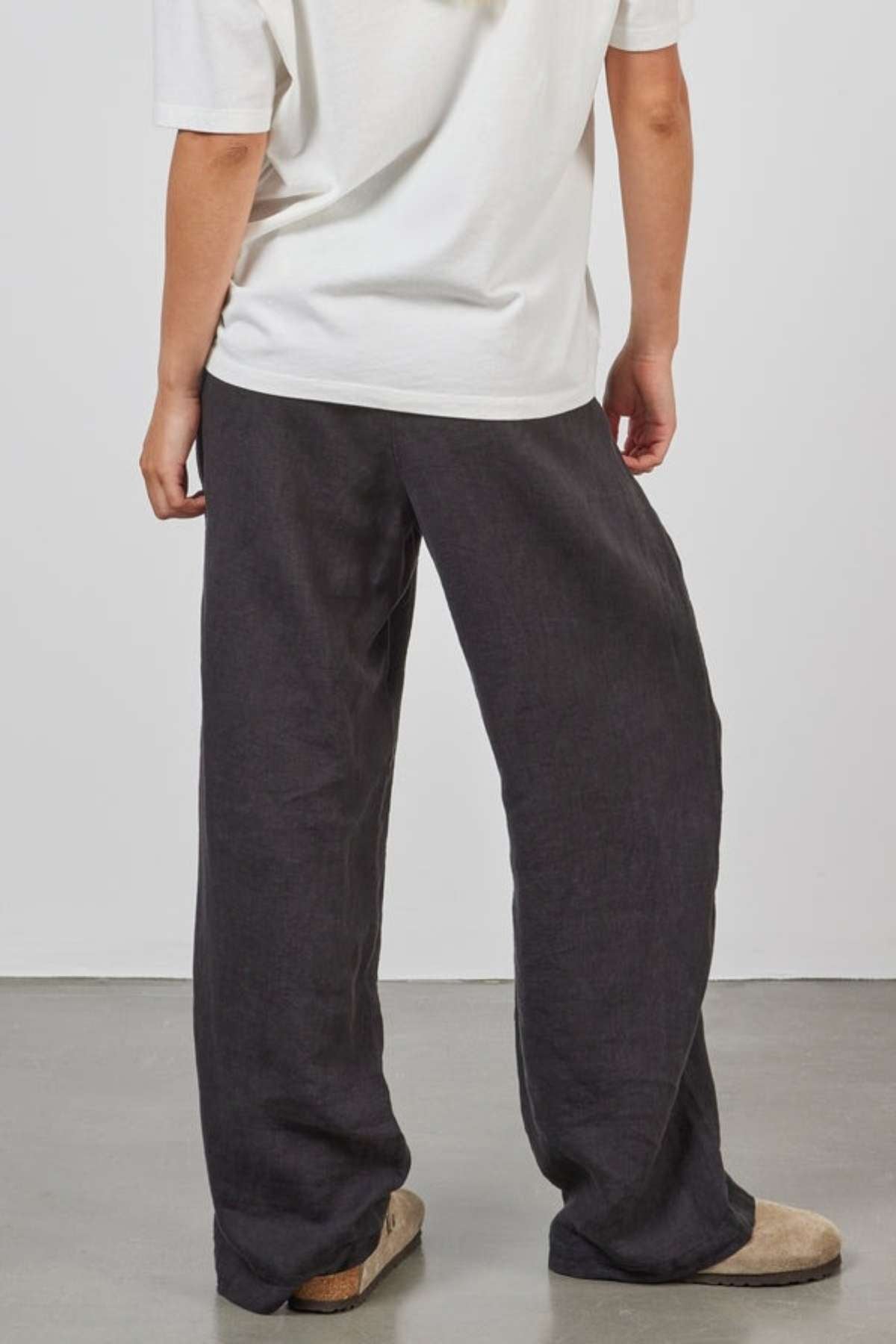 A person stands facing away from the camera, wearing a loose-fitting white t-shirt, Academy Women Riviera Linen Pants in black, and beige shoes. The background is plain and gray.