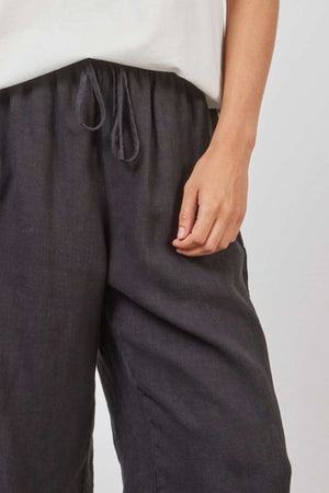 A person wearing a white top and dark Academy Women Riviera Linen Pant. Only their torso and part of their right arm are visible. Their right arm is relaxed by their side against a plain background, showcasing the relaxed wide leg design for a lightweight and breathable feel.