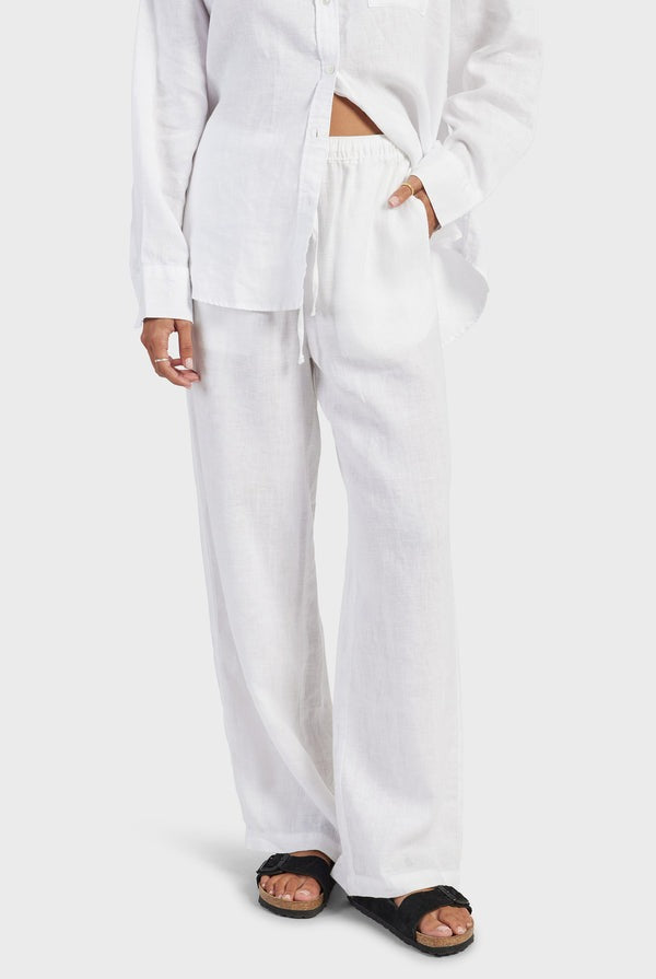 A person stands against a plain background, wearing a loose-fitting, button-up white shirt and matching white Academy Women Riviera Linen Pants with pockets. They have one hand in a pocket, and black sandals are visible on their feet. The outfit offers a comfortable fit, perfect for staying lightweight and breathable all day.