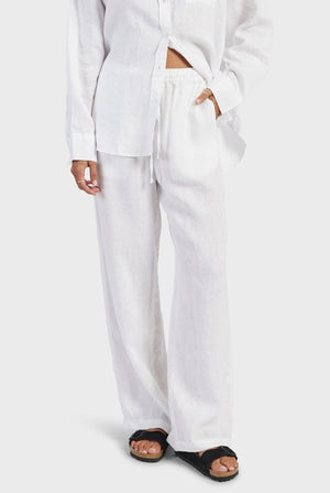 A person stands against a plain background, wearing a loose-fitting, button-up white shirt and matching white Academy Women Riviera Linen Pants with pockets. They have one hand in a pocket, and black sandals are visible on their feet. The outfit offers a comfortable fit, perfect for staying lightweight and breathable all day.