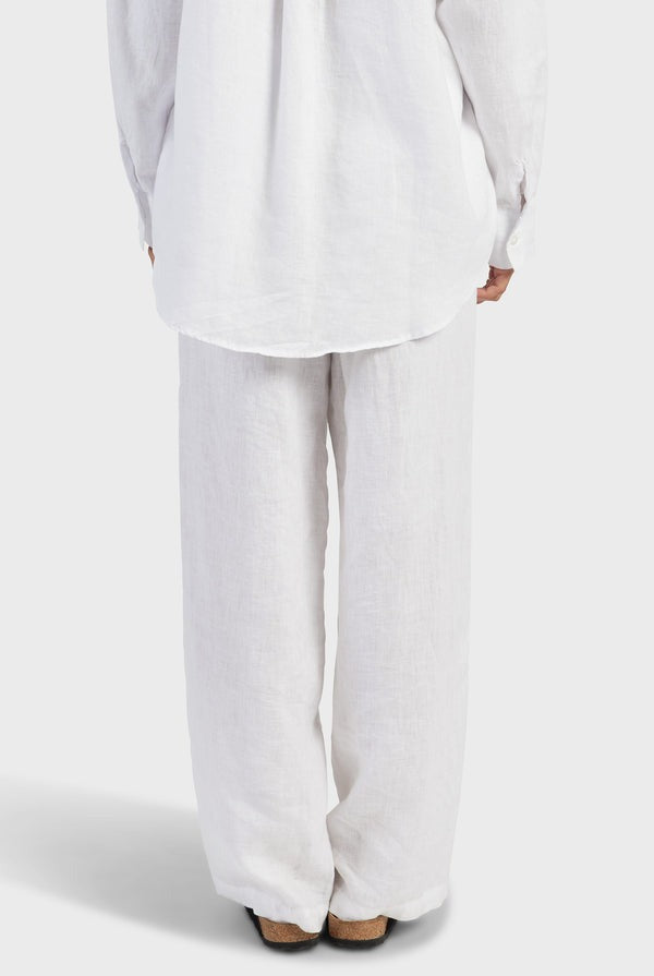 The image depicts a person standing with their back to the camera, dressed in a white, long-sleeved shirt and the Academy Women Riviera Linen Pant that offers a comfortable fit. The lightweight and breathable fabric seems to be linen. The background is plain and white.
