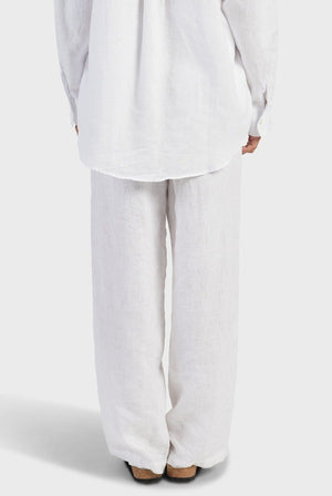 The image depicts a person standing with their back to the camera, dressed in a white, long-sleeved shirt and the Academy Women Riviera Linen Pant that offers a comfortable fit. The lightweight and breathable fabric seems to be linen. The background is plain and white.