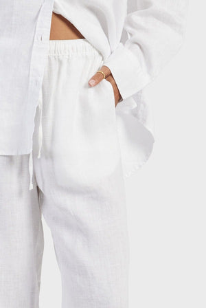 A person wearing a white long-sleeve button-up shirt and Academy Women Riviera Linen Pants stands with one hand casually in their pocket. The lightweight and breathable outfit appears comfortable and relaxed against a plain background.