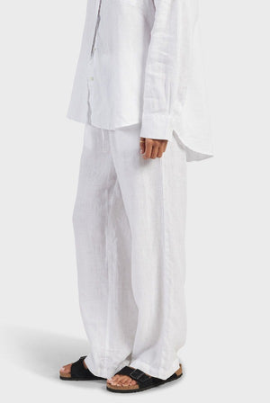 A person is wearing a white, long-sleeved button-down shirt and Academy Women Riviera Linen Pants, renowned for their lightweight and breathable fabric, paired with black sandals. The photo is cropped to showcase the comfortable fit of the outfit from the shoulders down against a plain white background.