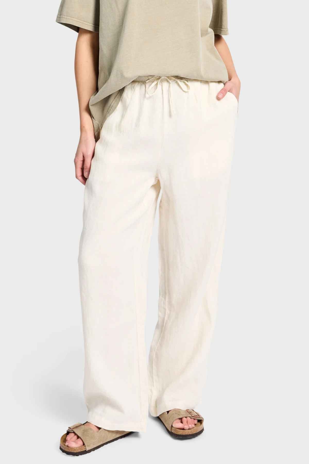 A person is wearing a beige T-shirt and the Academy Women Riviera Linen Pant, which features loose, wide-leg styling with an elastic waist, while casually tucking their hands in the pockets. They're also sporting beige sandals.