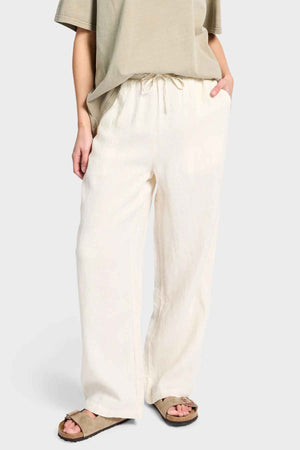 A person is wearing a beige T-shirt and the Academy Women Riviera Linen Pant, which features loose, wide-leg styling with an elastic waist, while casually tucking their hands in the pockets. They're also sporting beige sandals.