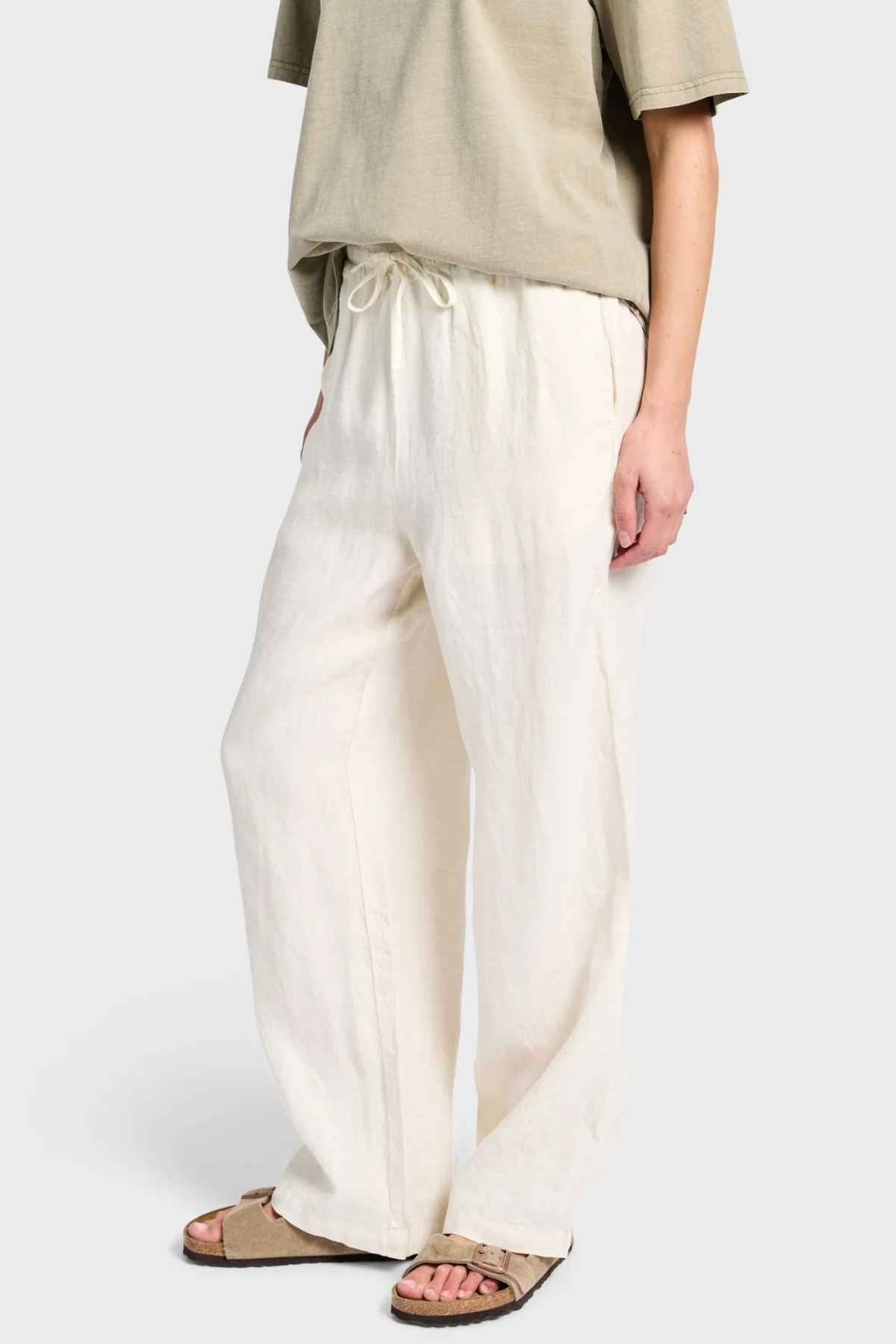 A person wearing a beige T-shirt and the Academy Women Riviera Linen Pant in cream, featuring an elastic waistband, stands against a plain background. They appear relaxed with one hand in their pocket, while wearing brown sandals.