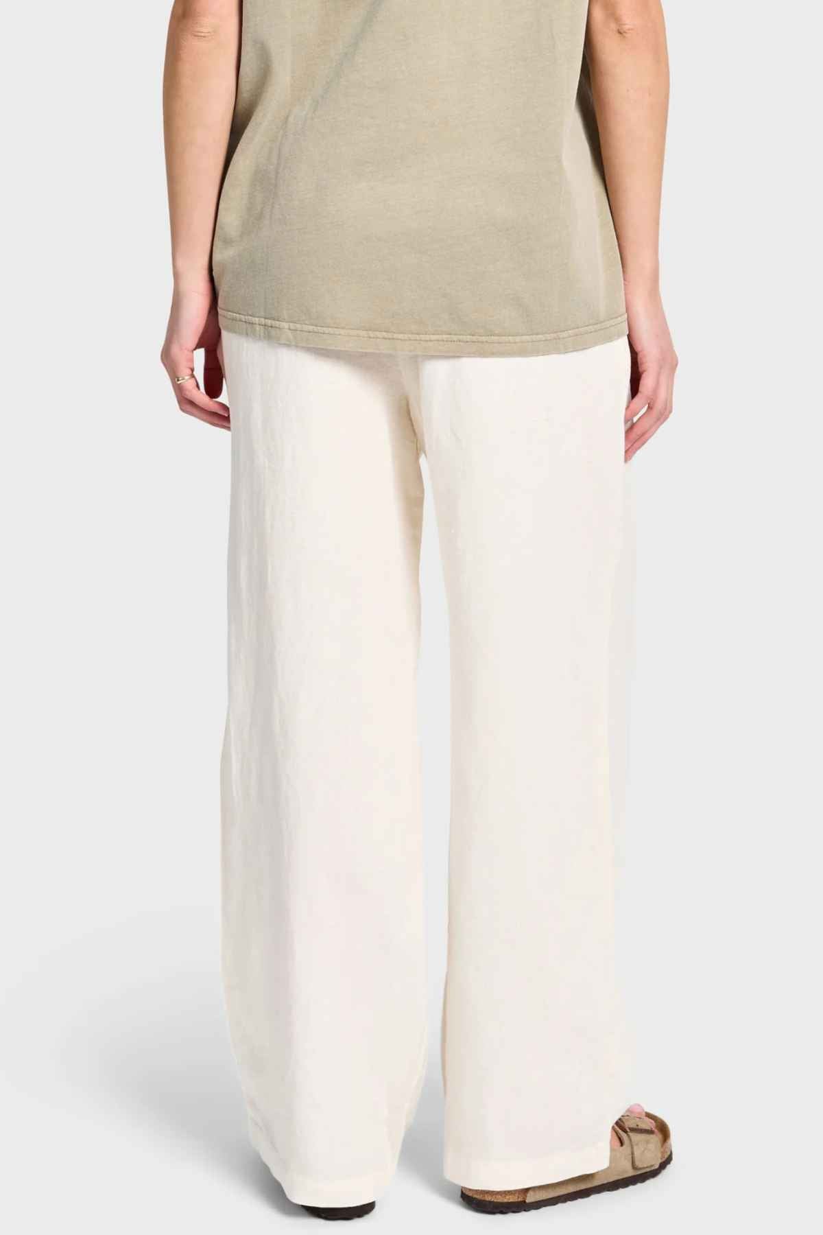 Rear view of a person in an olive green shirt and the Academy Women Riviera Linen Pant, designed with a wide-leg fit and elastic waist, standing against a plain background. The person is also wearing one sandal.