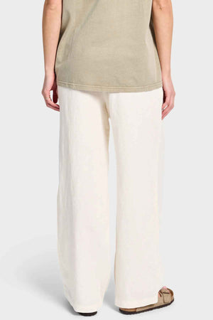 Rear view of a person in an olive green shirt and the Academy Women Riviera Linen Pant, designed with a wide-leg fit and elastic waist, standing against a plain background. The person is also wearing one sandal.