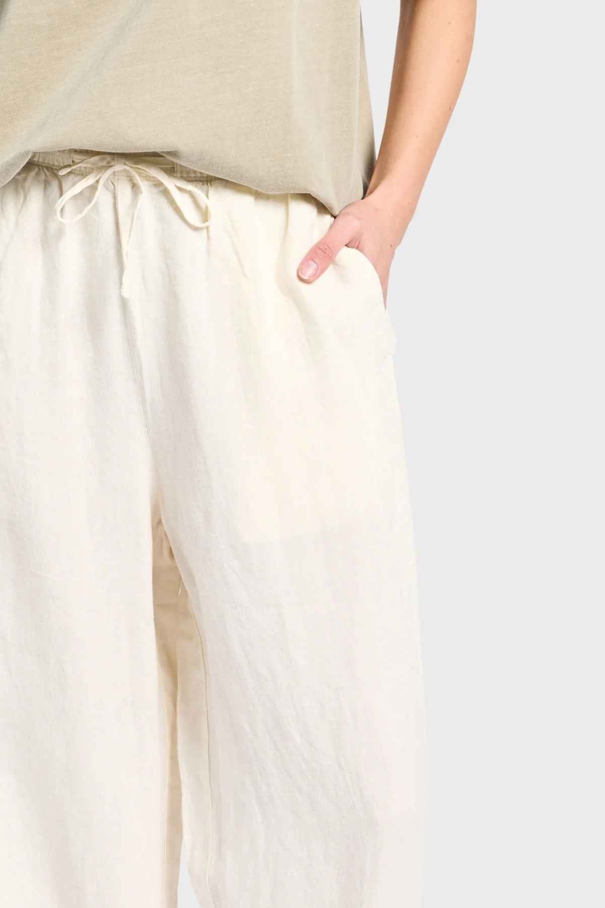 Someone wearing a beige top and the Academy Women Riviera Linen Pant, which are loose-fitting and wide-leg with an elastic waist, stands with one hand in a pocket against a plain light gray background.