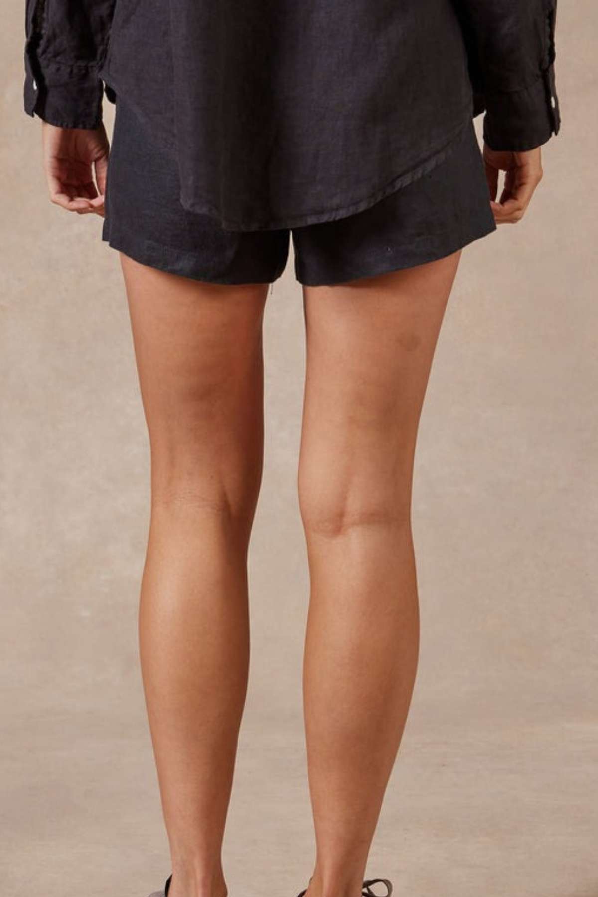 A person is standing with their back to the camera, wearing a black shirt and Academy Women Riviera Linen Short, which features an elastic waist and side entry pockets. The background is a plain, light-colored surface.