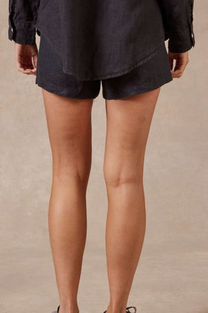 A person is standing with their back to the camera, wearing a black shirt and Academy Women Riviera Linen Short, which features an elastic waist and side entry pockets. The background is a plain, light-colored surface.