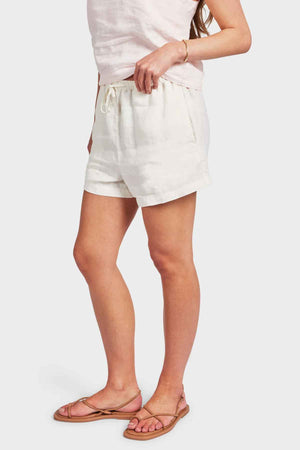 A person wearing a light-colored sleeveless top and Academy Women Riviera Linen Shorts, which feature side entry pockets and an elastic waistband, pairs them with tan sandals and a bracelet. They stand against a plain white background, effortlessly showcasing relaxed yet stylish summer attire.
