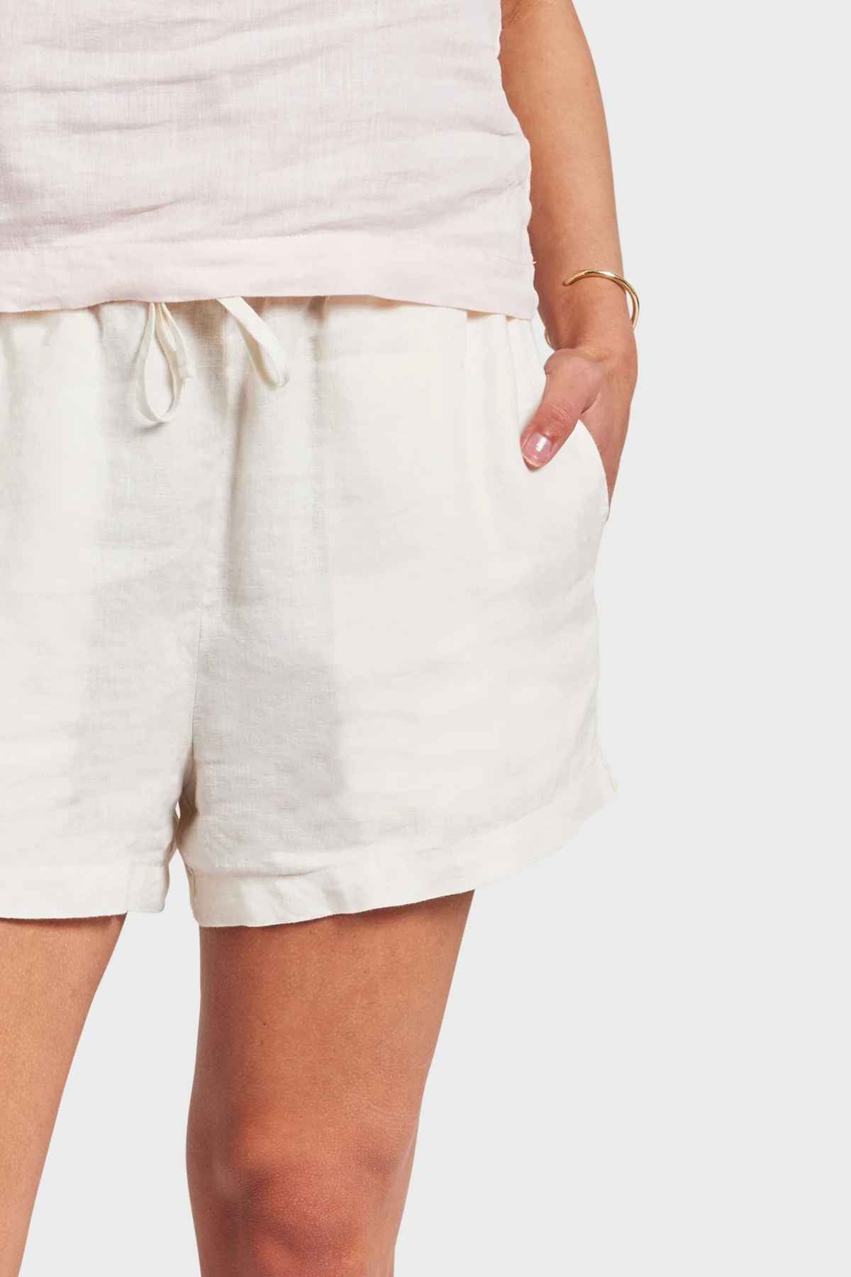 A person stands casually, dressed in a white linen top paired with the Academy Women Riviera Linen Shorts, featuring an elastic waistband and relaxed side entry pockets. The outfit highlights the texture of the fabric, while a simple bracelet on their wrist adds a touch of subtle elegance.