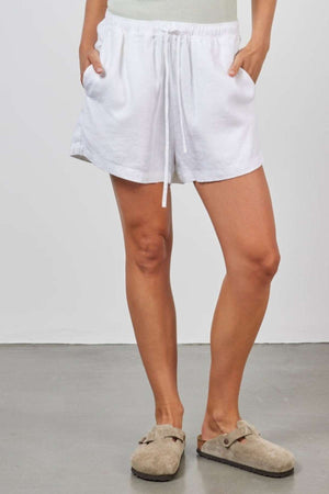 A person is standing on a gray floor wearing the Academy Women Riviera Linen Short with a drawstring and a light-colored top. They are also wearing beige slip-on shoes with dark socks. The enzyme-washed linen fabric of the shorts enhances the relaxed leg look, while their hands rest comfortably in the pockets.