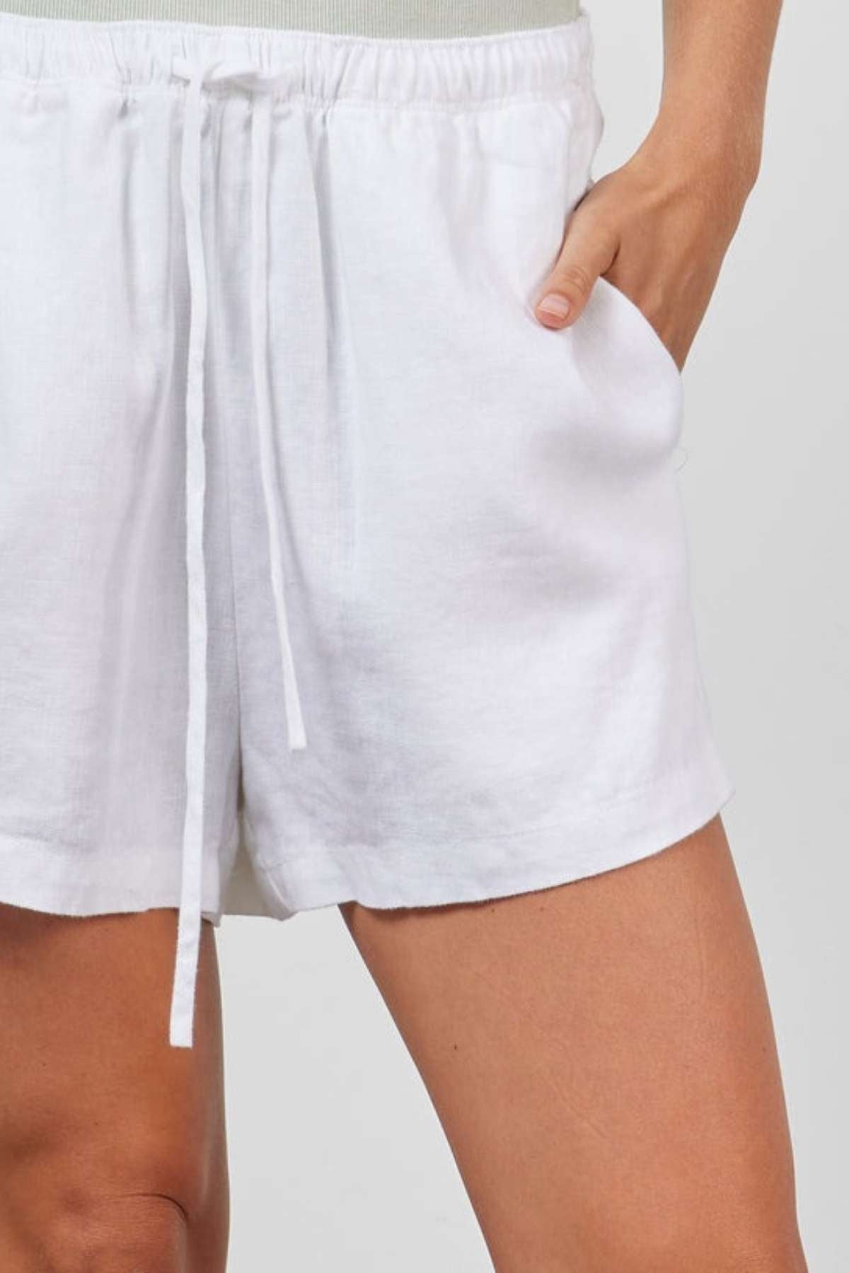 A person is depicted from the waist down wearing an Academy Women Riviera Linen Short, which features an elastic waistband and drawstring. Made from enzyme-washed linen, these shorts exhibit a relaxed leg fit. The individual's right hand is casually tucked into the pocket of the shorts against a plain, light-colored background.
