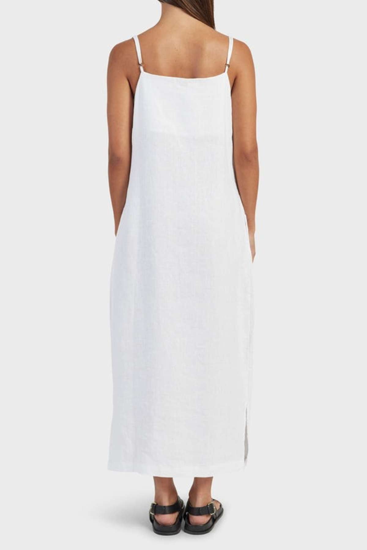 A person with long hair faces away in an essential summer piece: the Academy Women Essential Linen Slip Dress, a white sleeveless dress with thin straps. Paired with black sandals, it's perfect against the plain gray backdrop.