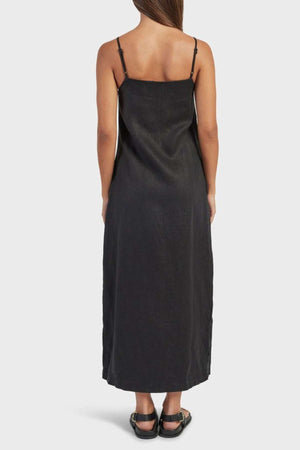 A woman stands facing away in the Academy Women Essential Linen Slip Dress—sleeveless, ankle-length, with adjustable straps and side splits—paired with black sandals. Her long brown hair is down against a plain white background.