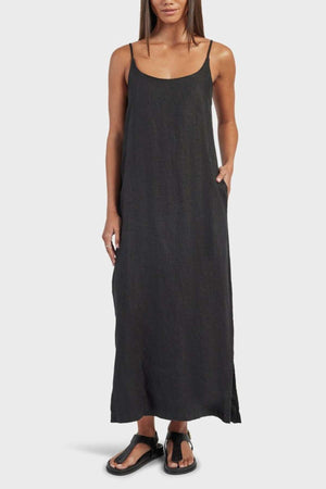 A woman wears the Academy Women Essential Linen Slip Dress, a loose black linen style with side splits and adjustable straps. She stands facing forward with one hand in her pocket, paired with black sandals against a plain light background.