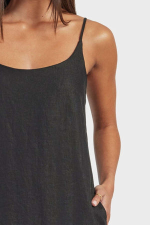 A woman wearing the Academy Women Essential Linen Slip Dress—sleeveless, black linen with adjustable straps and subtle side splits—stands against a light gray background, one hand in her pocket and her hair down.