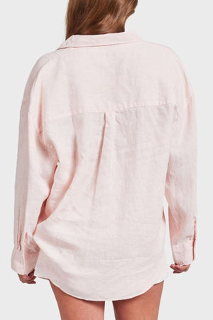 A person with long hair is wearing an oversized, light pink Academy Women Hampton Linen Shirt. The image captures the back view, showcasing mother-of-pearl buttons and a casual style against a plain background.