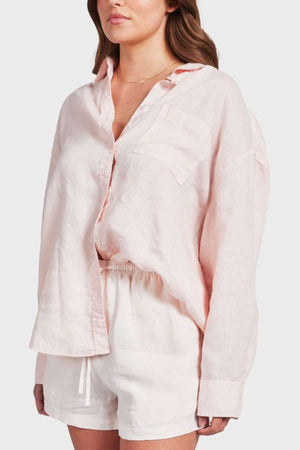A person wears an Academy Women Hampton Linen Shirt in light pink, featuring elegant mother-of-pearl buttons and a single pocket, paired with cream-colored shorts. The slightly oversized, garment-dyed shirt is tucked in neatly against a plain white background.
