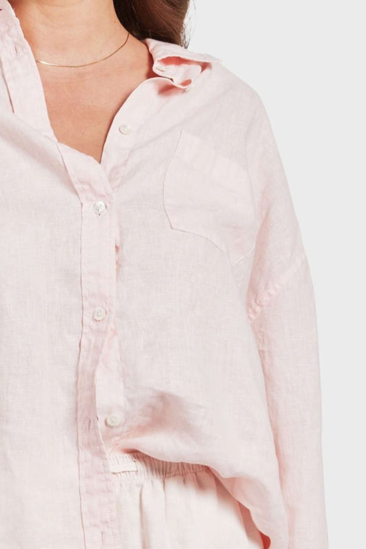A woman wears the Academy Women Hampton Linen Shirt in light pink with a chest pocket and a thin necklace, shown from shoulders to waist against a plain background for an effortlessly chic look.