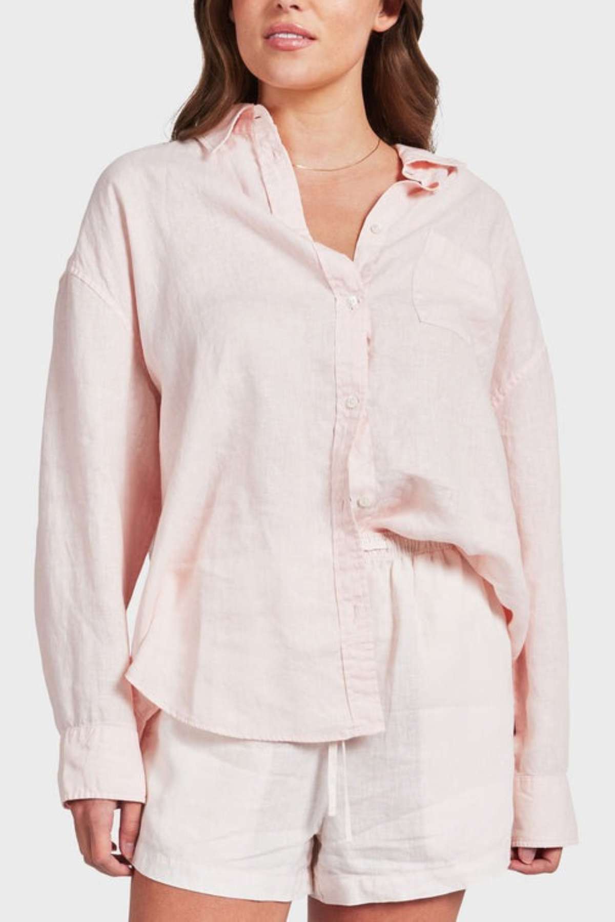 A woman in a light pink Academy Women Hampton Linen Shirt with mother-of-pearl buttons and long sleeves, paired with white shorts. The shirt is partially tucked in against a plain gray background.