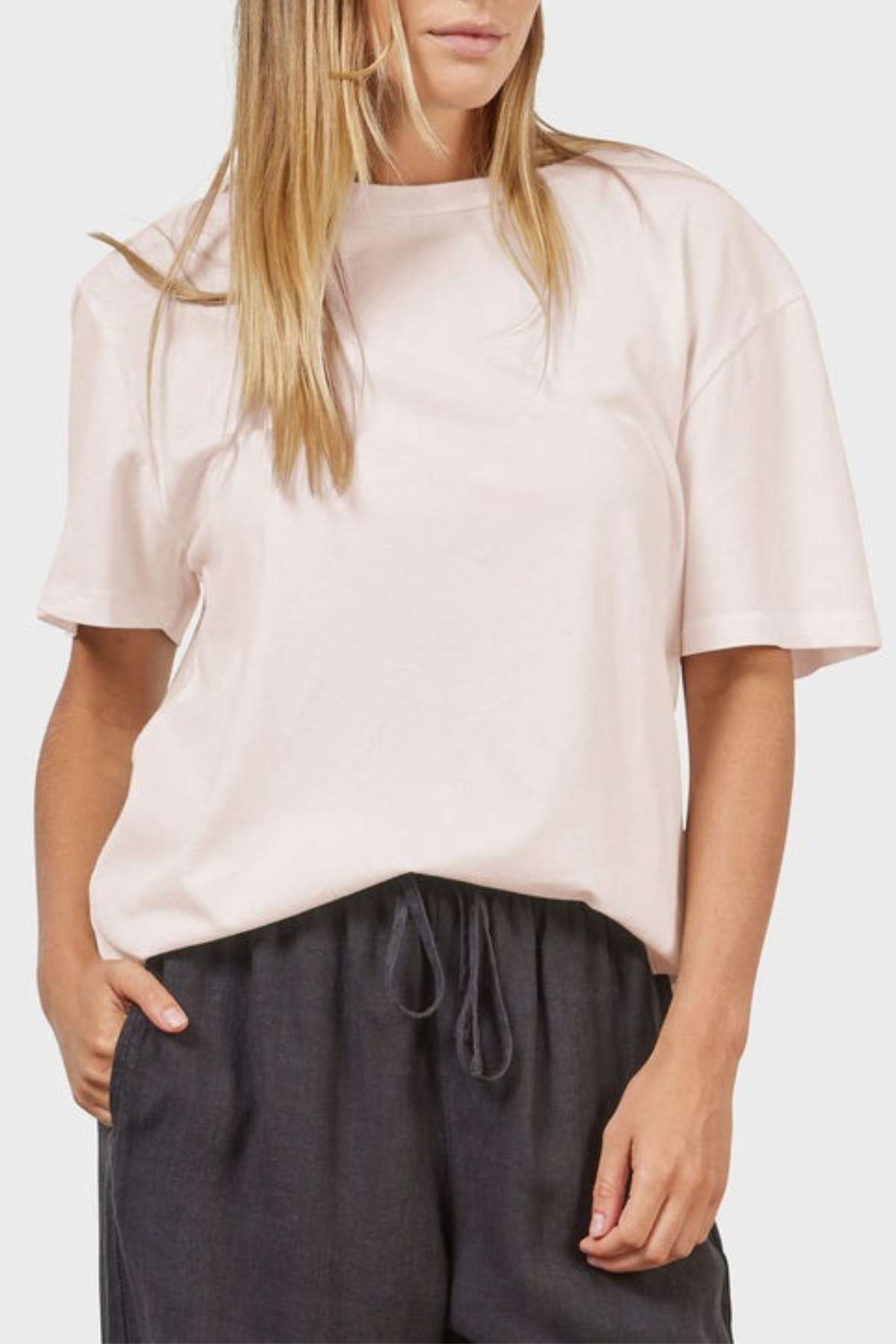With long hair, they wear an oversized Academy Women Jimmy Boyfriend Tee in light pink, paired with dark gray drawstring pants. The tee features a relaxed fit crafted from premium sanded jersey, as they stand against a plain background with one hand in their pocket.