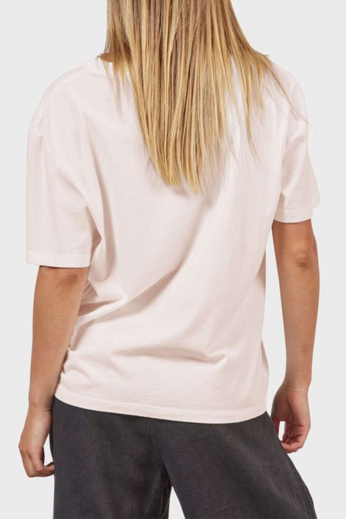 A person with long, blonde hair wears an Academy Women Jimmy Boyfriend Tee in crisp white, paired with dark pants. They stand back to the camera against a neutral background.